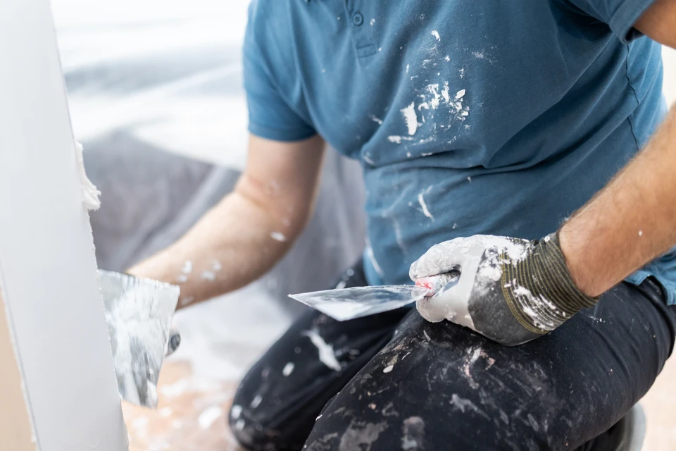 Photographie d'un artisan en tenue professionnelle discutant avec un client dans un intérieur en cours de rénovation, montrant plusieurs corps de métier en arrière-plan (menuisier travaillant sur une fenêtre, peintre, carreleur), illustrant la coordination et l'accompagnement global d'un projet de rénovation.